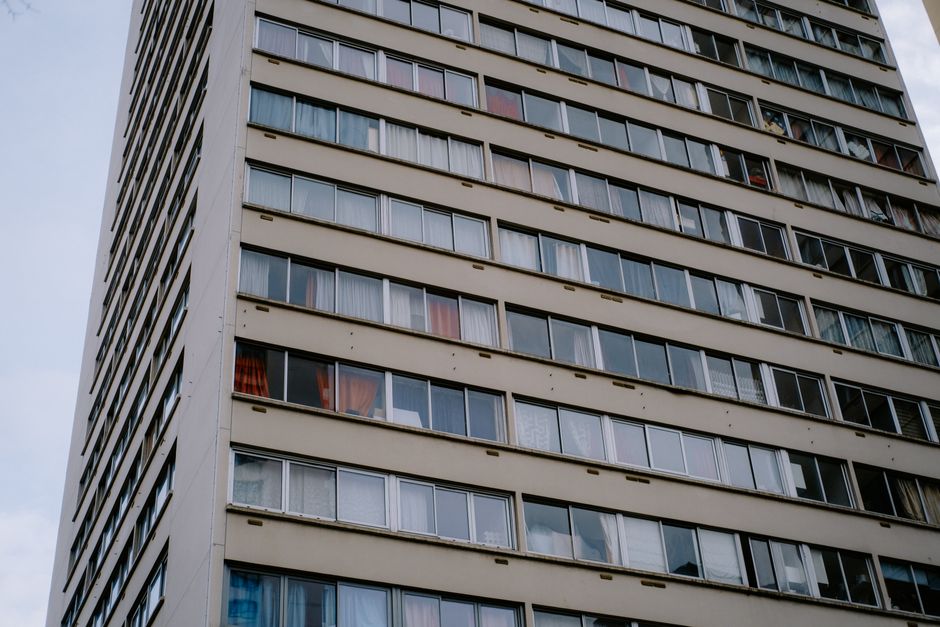A high-rise residential building with multiple floors, each featuring large windows, some of which have partially open curtains or blinds. The exterior facade of the building is light-colored with hor